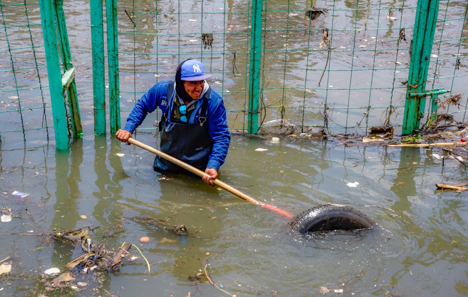 A man in a blue jacket and a blue New York Yankees cap is standing knee-deep in floodwater on Tottenham High Road, using a long-handled tool to remove debris from the submerged area near a green metal