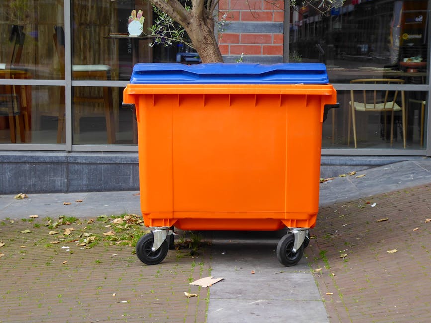 An outdoor scene featuring an orange waste bin with a blue lid and black wheels positioned on a paved sidewalk in front of a commercial building with large glass windows. The pavement around the bin h